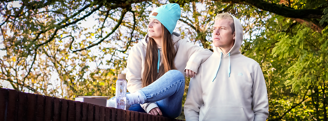 Zwei Personen tragen den PUEBLO Hoodie, im Hintergrund ist ein Ast mit Laub. Im Vordergrund steht die PUEBLO Glasflasche und die PUEBLO Lunchbox.